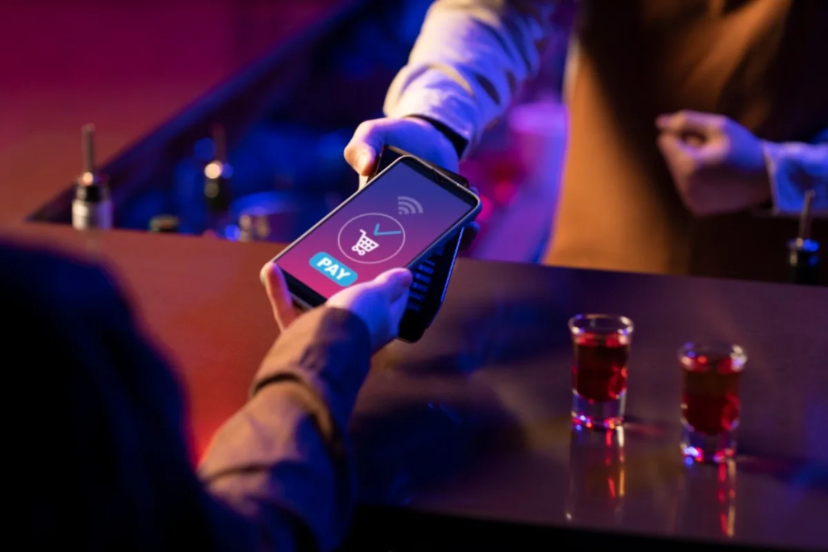 Contactless payment at a bar: smartphone tapped to a handheld card terminal, purple nightlife lighting